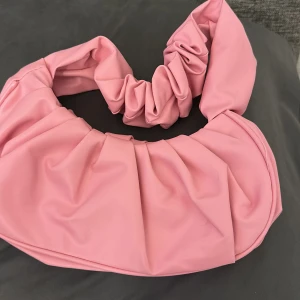 Rosa axelväska med scrunchie-handtag - Säljer en superfin rosa axelväska med unikt scrunchie-inspirerat handtag. Väskan har en mjuk, rundad form och stängs med magnetknapp. Perfekt för dig som vill ha en trendig accessoar som sticker ut.