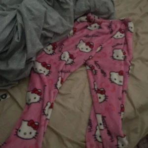 Hello Kitty mjukisbyxor i rosa fleece - Supergulliga rosa mjukisbyxor med Hello Kitty-motiv och resår i midjan. Byxorna är i mjuk fleece och har ett lekfullt mönster med Hello Kitty-ansikten och röda rosetter över hela tyget.