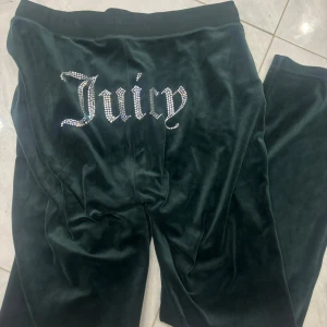 Mörkgröna velourbyxor från Juicy Couture - Mörkgröna mjukisbyxor i velour från Juicy Couture med glittrande stenar som bildar texten 'Juicy' på baksidan. Perfekta för en avslappnad och trendig look.