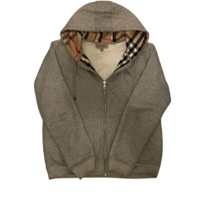 Grå hoodie från Burberry - Säljer en stilren grå hoodie från Burberry med klassiskt rutigt mönster i huvan. Tröjan har dragkedja och är tillverkad i mjuk bomull. Perfekt för en avslappnad look. Stl. L men sitter mer som en M. Påse och taggar medföljer