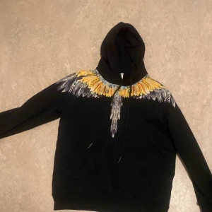 Svart hoodie med fjädermönster från Marcelo Burlon - Säljer en svart hoodie från Marcelo Burlon med färgglatt fjädermönster i gult, lila och vitt över axlar och bröst. Tröjan har huva med dragsko och klassisk passform. Perfekt statement-plagg för dig som gillar unika detaljer. Priset kan diskuteras 