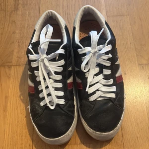 Svarta sneakers från Moncler med vita och röda detaljer - Säljer ett par svarta Moncler sneakers med vita snören och vit sula. Skorna har vita och röda ränder på sidan samt Moncler-logga. Insidan är ljus med perforerad innersula och rund tå. Perfekta för dig som gillar stilrena och sportiga skor. 