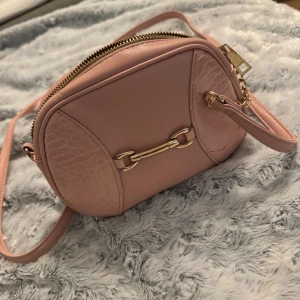 Ljusrosa axelväska med gulddetaljer - Säljer en söt ljusrosa axelväska med guldfärgad kedja och dekorativ metallspänne framtill. Väskan har dragkedja upptill och detaljer i krokopräglat mönster på sidorna. Perfekt storlek för det viktigaste och har justerbar axelrem.