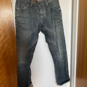 Blå/grå jeans från BOSS Orange - Säljer ett par blå jeans från BOSS Orange med orangea kontrastsömmar och knäppning med knappar. Jeansen har regular fit, klassiska bakfickor och en snygg läderpatch med logga bak i midjan.