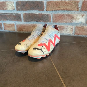 Vit och röda Puma fotbollsskor - Säljer ett par vita fotbollsskor från Puma med röda detaljer och svarta Puma-loggor. Skorna har snörning och platt sula med dobbar, perfekt för spel på gräsplan. Snygg och sportig design med stickad ovandel för extra komfort.