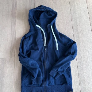 Mörkblå hoodie med dragkedja från Intimissimi - Säljer en mörkblå hoodie från Intimissimi med dragkedja och huva. Tröjan har långa ärmar, två fickor framtill och vita snören vid huvan. Perfekt för en avslappnad stil.