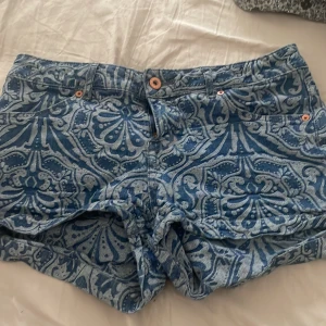 LÅG MIDJADE Blå mönstrade jeansshorts från Divided - Säljer ett par blå jeansshorts från Divided med ett unikt, vitt paisleymönster. Shortsen har klassisk femficksdesign, knapp och dragkedja framtill samt bälteshällor. Perfekta för sommaren och ger en cool, avslappnad look.