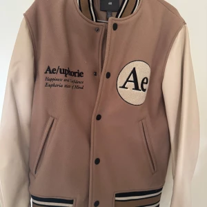 Beige och vit varsityjacka från Ae/uphorie - Säljer en beige och vit varsityjacka från Ae/uphorie med broderad text och emblem på bröstet samt stor logga på ryggen. Jackan har svarta knappar, randiga muddar och två fickor framtill. Perfekt för en sportig och trendig look.