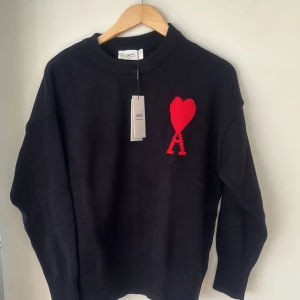 Svart stickad tröja från Ami Paris - Svart stickad tröja från Ami Paris med rund halsringning och röd broderad logga i form av ett hjärta och bokstaven A på bröstet. Tröjan har långa ärmar och ribbade muddar vid hals, ärmslut och nederkant.