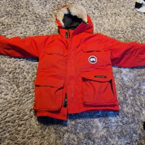 Röd vinterjacka från Canada Goose - Röd vinterjacka från Canada Goose med huva fodrad i fuskpäls. Jackan har flera fickor med dragkedja och knappar samt tydlig logga på bröstet och ärmen. Perfekt för kalla dagar och riktigt varm.