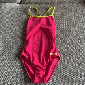 Rosa baddräkt från Arena med gula detaljer - Säljer en sportig rosa baddräkt från Arena med tunna gula axelband och logga framtill. Baddräkten har öppen rygg och är perfekt för simträning eller bad. Snygg och färgstark design!