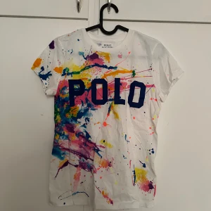 Vit t-shirt med färgstänk från Polo Ralph Lauren - Säljer en vit t-shirt från Polo Ralph Lauren med stora blå bokstäver 'POLO' på bröstet och färgglada färgstänk i rosa, gul, blå, lila och grön. T-shirten har rund hals och korta ärmar. Perfekt för dig som gillar unika och färgglada plagg!