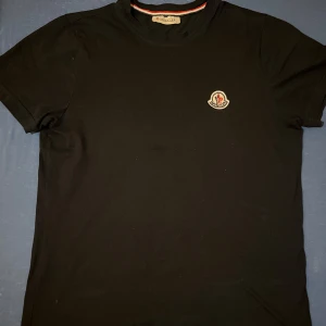 Moncler T-shirt Svart - Säljer en svart Moncler t-shirt i storlek S. Klassisk modell med Monclers logga på bröstet. Mjuk och högkvalitativ bomull, normal passform. Väldigt sparsamt använd – inga fläckar, hål eller slitningar. Tyvärr finns inget kvitto kvar då det var en present, men äktheten kan garanteras på material, detaljer och passform. Enkel att styla och passar både till vardag och fest!