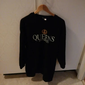 Svart sweatshirt med tryck 'QUEENS NEW YORK CITY' - Svart sweatshirt med tryck i vitt och grönt där det står 'QUEENS NEW YORK CITY' samt ett litet emblem ovanför texten. Tröjan har rund hals och långa ärmar. Perfekt för en avslappnad streetstil.