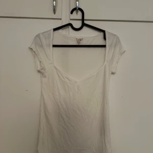 Vit topp med spetskant - Söt vit topp med djup rundad ringning och spetsdetalj längs kanten. Kortärmad och i mjukt material, perfekt för sommardagar. Enkel och stilren design som passar till mycket.