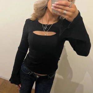 Svart ribbad långärmad topp med cut-out - Trendig svart ribbad topp med lång ärm och snygg cut-out detalj vid bröstet. Toppen har en figurnära passform och är perfekt för att lyfta din outfit med en cool touch. Snyggt att matcha med jeans eller kjol.
