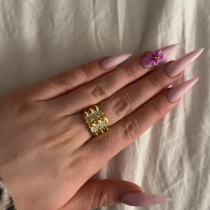 Bred guldfärgad ring med stenar och nitar - Säljer en bred guldfärgad ring med tre rader av gnistrande stenar. Ringen har en modern och chunky design som sticker ut och passar perfekt som statement-smycke. Levereras i en röd presentask med rosett.