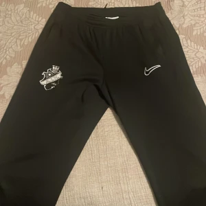 Svarta AIK träningsbyxor från Nike - Svarta träningsbyxor från Nike med AIK-logga på ena benet och Nike Swoosh på andra. Byxorna är i Dri-Fit material som andas och har en sportig, smal passform. Perfekta för fotbollsträning eller annan sport.