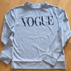 Ljusgrå sweatshirt med VOGUE-tryck - Säljer en ljusgrå sweatshirt med svart VOGUE-tryck på ryggen. Tröjan har lång ärm och en högre krage. Perfekt för dig som gillar stilrena plagg med statement-tryck.