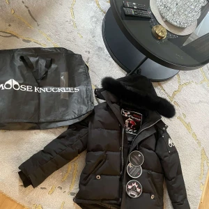 Svart pufferjacka från Moose Knuckles - Säljer en svart pufferjacka från Moose Knuckles med avtagbar huva och snygg pälsdetalj. Jackan har dragkedja, knappar och två fickor framtill. Ikonisk logga på ärmen och quiltad design. Perfekt för kalla dagar. INKÖPT FÖR 11 tusen från johnells!!!