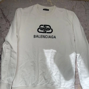 Vit sweatshirt från Balenciaga - Säljer en vit sweatshirt från Balenciaga med svart logga och tryck framtill. Tröjan har rund halsringning och långa ärmar. Tillverkad i bomull och passar perfekt för en stilren och trendig look. L