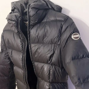 Svart dunjacka från Colmar - Säljer en svart dunjacka från Colmar med huva och quiltad design. Jackan har lång ärm, dragkedja framtill och en klassisk Colmar-logga på ena ärmen. Perfekt för kyliga dagar och enkel att matcha med olika outfits. 