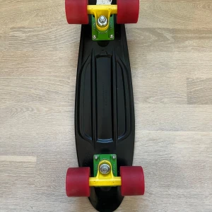 Penny Australia - Säljer en svart skateboard i plast med gröna och gula truckar samt röda hjul. Brädan har räfflad ovansida för bättre grepp och är kompakt i formen, perfekt för att cruisa runt i stan.