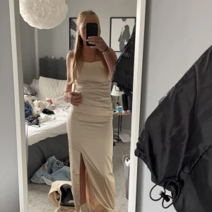 Champagnefärgad långklänning med slits - Elegant champagnefärgad långklänning med smala axelband och en stilren slits framtill. Klänningen har en öppen rygg med snörning som ger en sofistikerad look. Perfekt nu inför balen☺️🥂Köpt för 