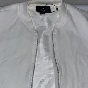 Vit bomberjacka från MAN - Snygg vit bomberjacka från MAN med dragkedja framtill. Jackan har en stilren design och är perfekt för en casual look. Den är långärmad och har en klassisk bomberstil.