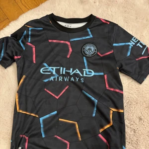 Svart fotbollströja från Manchester City - Säljer en svart fotbollströja från Manchester City med färgglada geometriska mönster i blått, rött och orange. Tröjan har korta ärmar och spelarens namn och nummer 'Haaland 9' på ryggen. Perfekt för fans som vill visa sitt stöd! DEN ÄR INTE ÄKTA