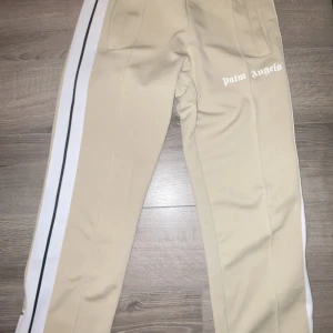 Palm Angels byxor  - Snygga beige byxor från Palm Angels med vita och svarta ränder längs sidorna. Byxorna har en dragkedja vid bensluten och är tillverkade i polyester. Perfekta för en stilren look.