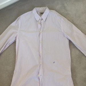 Randig skjorta från Acne - Säljer en stilren randig skjorta från Acne i ljuslila och vit. Skjortan har långa ärmar och knappar framtill. Den är broderad med ett  'A' på bröstet för att visa märket. Nya kostar acne skjortor kostar mellan 1500-5000kr