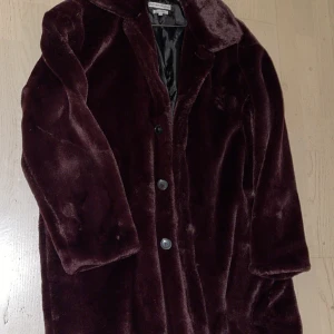 Vinröd/lila pälsjacka (fuskpäls) - Säljer en elegant vinröd/lila pälsjacka med knappar framtill. Jackan har en klassisk design med långa ärmar och en mjuk, lyxig känsla. Perfekt för att hålla sig varm och stilren under kyliga dagar.