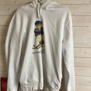 Vit hoodie från Ralph Lauren - Säljer en stilren vit hoodie från Ralph Lauren med en cool Polo Bear-design på framsidan. Perfekt för en avslappnad look. Hoodien har en klassisk passform med en bekväm huva och ribbade muddar.