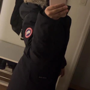 Svart vinterjacka med pälsluva - Säljer en svart vinterjacka från Canada Goose med pälsdetalj på huvan. Jackan har en klassisk design med lång ärm och är perfekt för kalla dagar. Den har en tydlig logotyp på ärmen.