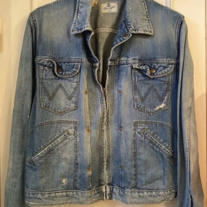 Blå jeansjacka från Wrangler - Säljer en klassisk blå jeansjacka från Wrangler. Jackan har en normal passform med långa ärmar och knappar framtill. Den har två bröstfickor med lock och två snedställda fickor nedtill. Perfekt för en avslappnad stil.