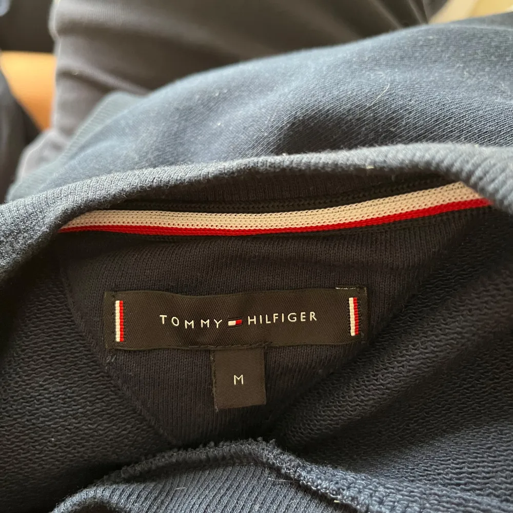 Säljer en stilren mörkblå tröja från Tommy Hilfiger med märkets logga på bröstet och text längs ärmarna. Tröjan har långa ärmar och en rund halsringning. Perfekt för en casual look. Modellen är 185 cm och väger 100 kg. . Neuletakit & Villapaidat.