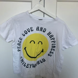 Vit t-shirt med smiley - Säljer en vit t-shirt från SmileyWorld men är köpt på Lindex med ett stort gult smileytryck och texten 'Peace Love and Happiness'. Är i stolek 158-164 men är liten för den storleken.