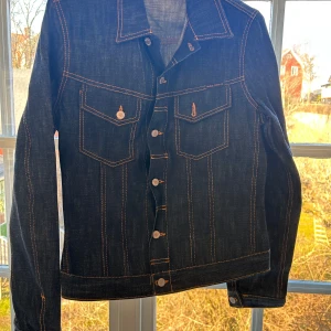Raw Denim Jeans Jacka från Nudie - Snygg mörkblå jeansjacka från Nudie med kontrasterande sömmar och knappar framtill. Jackan har två bröstfickor med lock och knapp, samt klassisk krage. Perfekt för en avslappnad stil.