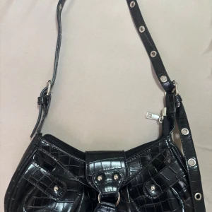 Svart axelväska med krokodilmönster - Snygg svart axelväska i syntet med krokodilmönster och justerbar axelrem. Väskan har dragkedja och dekorativa nitar på framsidan. Perfekt för att ge en edgy touch till din outfit.