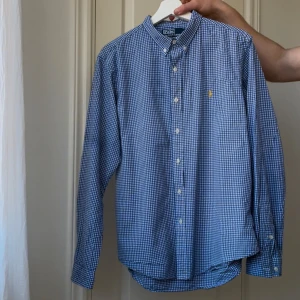 Polo Ralph Lauren skjorta långärmd - Säljer en stilren blå och vit rutig skjorta från Polo Ralph Lauren. Skjortan har en slim fit passform och är dekorerad med ett gult broderat märke på bröstet. Den har långa ärmar och knappar framtill. Perfekt för en klassisk look! Finns missfärgning,  märks knappt. Kan gå bort, har ej provat ta bort det ordentligt 