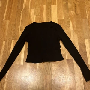 Svart ribbad topp med knyt i ryggen  - Säljer en svart ribbad topp med långa ärmar och en snygg knytning i ryggen. Perfekt för en stilren look. Toppen är croppad och är fin till många under delar.