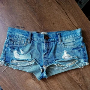 Blå jeansshorts med slitningar - Säljer ett par blå jeansshorts med coola slitningar och fransiga kanter. De har en knapp och dragkedja framtill samt fem fickor. Perfekta för en avslappnad sommarlook! Storlek UK 8 vilket motsvarar 34 i EU. | Midjemått ca 38 cm
