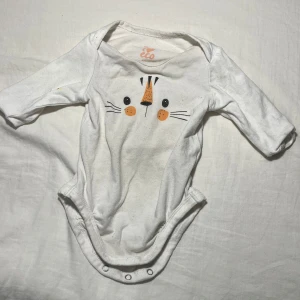 Vit långärmad body med kattmotiv från ICA - Söt vit långärmad body från ICA med ett gulligt kattansikte tryckt på framsidan. Tillverkad i ekologiskt material, perfekt för de minsta. Praktiska knappar i grenen för enkel på- och avklädning.