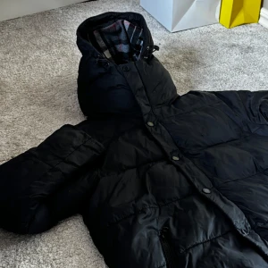 Svart dunjacka från burberry - Höj din stil denna säsong med en tidlös svart dunjacka från Burberry!   - Märke: Burberry - Färg: Svart - Material: Högkvalitativt dun för optimal värme och komfort - Funktioner: dragkedjor vid fickorna, en ficka inuti också.   Denna jacka är perfekt för både vardag och fest, och ger en lyxig känsla oavsett tillfälle. Burberrys klassiska design säkerställer att du alltid ser stilren ut.