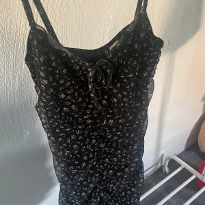 Svart blommig klänning med knyt - Snygg svart klänning med små blommor och knytning framtill. Perfekt för en somrig look. Klänningen har en smickrande passform och tunna axelband.