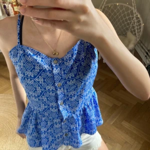 Blå blommig blus med knappar - Säljer en söt blå blus med blommigt mönster och knappar framtill. Den har smala axelband och en volangdetalj vid nederkanten. Perfekt för sommardagar! 💙