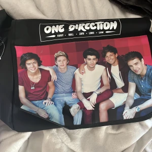 Svart axelväska med One Direction-tryck - Säljer en svart axelväska med ett stort tryck av One Direction på framsidan. Väskan har ett justerbart axelband och är perfekt för fans av bandet. Den har en praktisk storlek för att bära med sig det nödvändigaste.