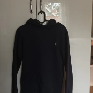 Blå hoodie från Ralph Lauren - Säljer en stilren mörkblå hoodie från Ralph Lauren med det klassiska logotypen broderad på bröstet. Perfekt för en avslappnad look. Den har en bekväm passform och en praktisk ficka framtill. Storlek XL i barn men passar S i herr. Passar mig bra som är 174 så allt mellan 170-175 