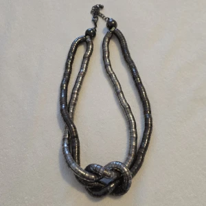 Dubbelradigt halsband i metallisk stil - Snyggt dubbelradigt halsband med metalliska skivor i en mörk silverfärg. Halsbandet har en unik knutdetalj framtill och justerbar kedja baktill för perfekt passform. Perfekt för att ge en edgy touch till din outfit.